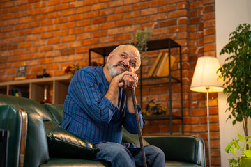 Old Indian man in kurta holding walking stick, sitting alone on sofa with helpless expression