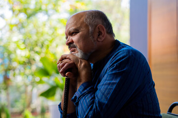 Old Indian man in kurta holding walking stick, sitting alone on sofa with helpless expression