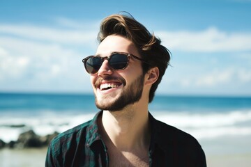 Man with a beard and sunglasses is smiling at the camera. He is wearing a plaid shirt and he is enjoying the beach