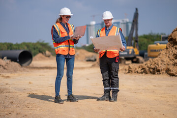 Construction surveyor or engineer working construction site blueprint and laptop for inspection detail of construction with heavy machinery excavator constructed hi-way road background.