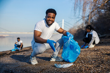 Portrait of interracial activist putting plastic and paper waste into trash bag with his environmentally conscious team at riverbank.