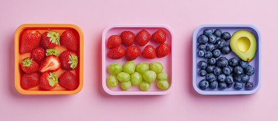 Colorful flat lay of three lunch boxes featuring red strawberries, green grapes, and dark blueberries arranged on a pastel pink background.