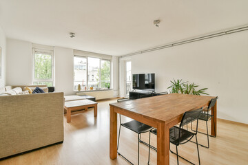A spacious and bright living room featuring a wooden dining table, minimalist decor, and a large window showcasing greenery outside.