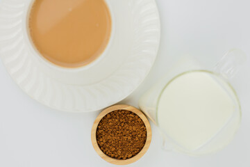 Close-up view from above: a white mug of coffee with milk standing on a saucer, next to it is a wooden bowl of instant ground coffee and a jug of milk on a white background. The concept of