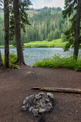 This serene lakeside campsite showcases a stone fire pit nestled under towering pine trees, with vibrant greenery and a calm body of water in the background, perfect for relaxation.