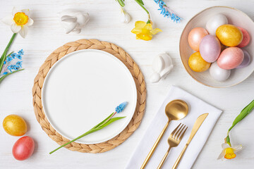 Top view of Easter table setting adorned with colorful spring flowers, white empty ceramic plate and painted eggs