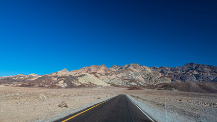 An enchanting road winds through a varied mountainous desert landscape, inviting travelers to explore the vast and beautiful terrains under a bright blue sky, symbolizing adventure and journey.