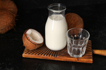 Wooden cutting board and bottle of tasty fresh coconut milk on black background, closeup