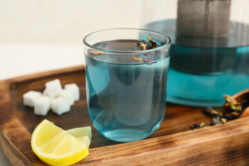Cup of butterfly pea flower tea, lemon piece and wooden tray on light background, closeup