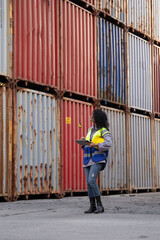 Female engineer standing at shipping container yard inspecting cargo delivering loading document on chart. Afro hair african woman in uniform checking import export container at logistic terminal dock
