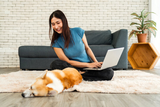 A young woman works on her laptop while her Corgi dog sleeps on a cozy rug. The scene captures remote work, pet companionship, work-life balance, and a relaxed home environment.