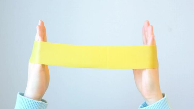 Female hands stretching a yellow fitness elastic band on gray background close-up.