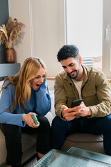 Excited young couple sharing a joyful moment while looking at a smartphone together in their cozy living room with laughter and smiles on their faces, enjoying their time together and creating lasting