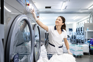 Smiling female worker holding laundry basket and operating washing machine in dry cleaning service.