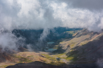 Aerial top view to green rocky hilly valley with two turquoise lakes under lush low clouds. Most...