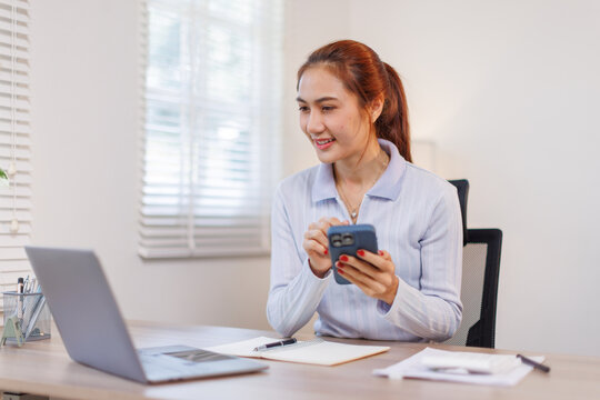 Serious charming asian woman using smartphone while working with laptop at home workplace.
