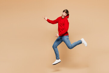 Full body side view musician singer young Caucasian man he wear red hoodie casual clothes jump high play air guitar isolated on pastel plain light beige background studio portrait. Lifestyle concept.