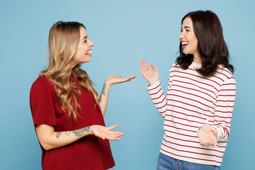 Side view young couple two friends women wear red casual clothes talk speak to each other spread hands together isolated on pastel plain light blue cyan background studio portrait. Lifestyle concept.
