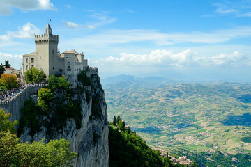 Naklejka premium A stunning view of a medieval castle perched on a rocky cliff, surrounded by lush green hills and valleys under a bright blue sky with fluffy clouds.