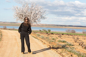 Una mujer vestida de negro con gafas de sol posa en un camino de tierra junto a un árbol en flor,...