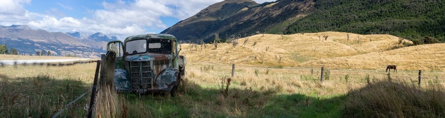 A rusted green truck sits abandoned in a golden field, framed by wooden fence posts. A horse grazes nearby, with lush green mountains rising in the background