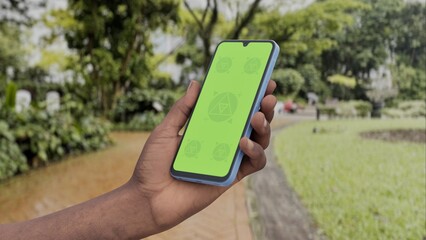 GREEN SCREEN CHROMA KEY Person holding a smartphone with a green screen in a lush garden environment, surrounded by greenery and a scenic path during daylight