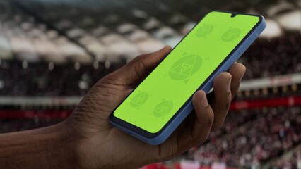 GREEN SCREEN CHROMA KEY A hand holding a phone with a green screen at a stadium during a football match. Ideal mockup for sports, betting, or event apps