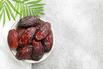 Many tasty dried dates in bowl and leaves on gray textured table, top view. Space for text