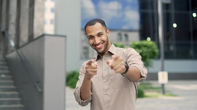 Handsome young adult man pointing fingers of both hands looks at camera showing joyful gesture standing on street near office building. Mixed race hr businessman in shirt smile pleased happy choosing