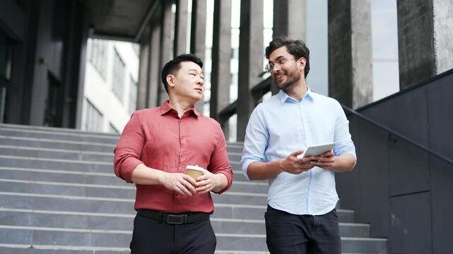 Two male colleagues walking down stairs outside office building discussing business, one holding coffee cup and other using tablet. Collaboration, and professional atmosphere in corporate environment