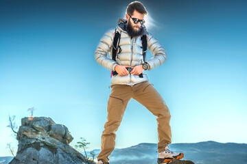 Confident traveler in the mountains. A bearded man in a stylish outdoor outfit, sunglasses, and a backpack stands on a rock, adjusting his gear under the bright blue sky