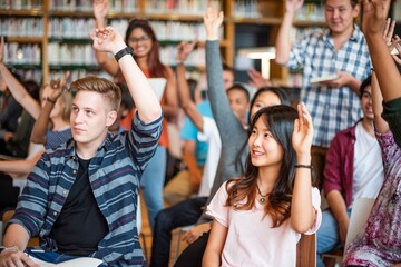 Diverse group of students in a library raising hands. Engaged students, mixed ethnicities, actively participating in a learning environment. Diverse students in library, education concept.
