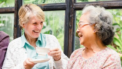 Two elderly women laughing and enjoying tea together by a window. Elderly women sharing a joyful moment. Smiling elderly women with tea cups. Diverse elderly friends enjoying tea at the table.
