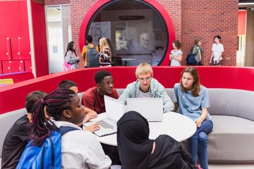 Diverse group of students collaborating in a modern study area. Students with laptops, engaged in teamwork. Bright, open space with red accents. Diverse students studying in school.