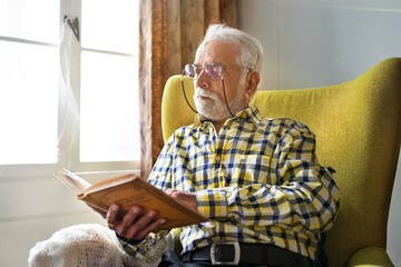 Fototapeta na wymiar Senior man with glasses reading a book in a yellow chair by a window. Wearing a plaid shirt, he appears focused and relaxed in a cozy setting. Senior man reading a book, sitting by window in chair.