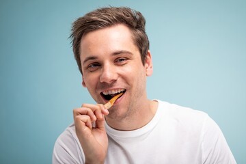 Man eating a fry, smiling, against a blue background. Casual, happy expression. Enjoying a snack, wearing a white shirt, light blue backdrop. Man cleaning teeth, dental hygiene.