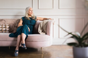 Elegant older woman wearing dress, sits on couch. The older woman looks thoughtful. Elegace old...