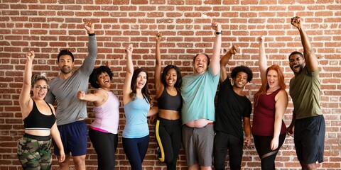 Diverse group of people standing in front of a brick wall, smiling and raising their arms in celebration. Diverse men and women in fitness wear raise their hand together for unity. Fitness people