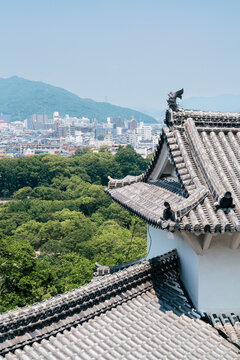 The ornate rooftop of Himeji Castle overlooks a lush green landscape and a distant city skyline, showcasing the contrast between traditional architecture and modern urban life.
