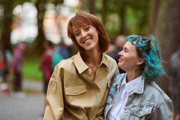 Fototapeta premium Young women standing close in park. One with brown hair, wearing beige jacket other with blue hair, denim jacket. Both smiling. Blur of trees, people in background