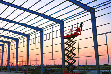 Silhouette of welder worker on electric scaffolding is welding metal structure on factory building wall framework in construction site area at sunset time