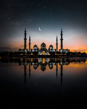 View of al zayed mosque illuminated at night with reflection in water and crescent moon, Abu Dhabi, United Arab Emirates.