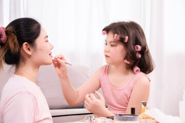 Cheerful young girl with pink curlers in her hair shares loving moment with her mother, enjoying fun and affectionate beauty routine at home. Take turns applying makeup, family warmth, spending time.