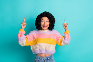 Portrait of a cheerful young woman pointing up with both hands wearing a colorful sweater against a...
