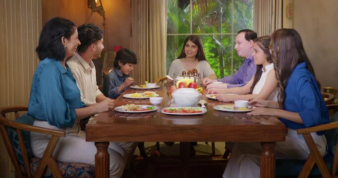 Indian happy family group with cute kid child sitting around dining table chair enjoy night dinner adult dad teen girl young boy talk mom lady serve fresh salad eat tasty food have fun joy spend time