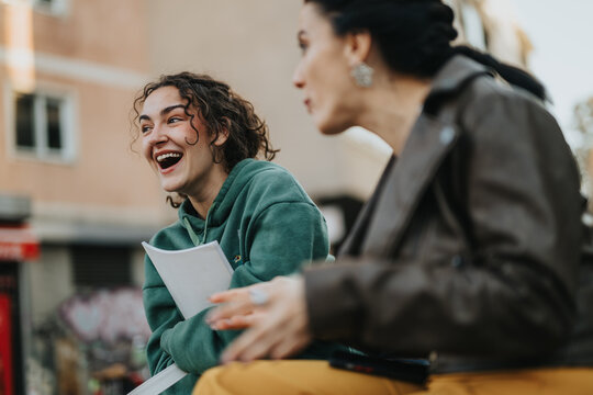 Two young women enjoy a fun conversation outside, sharing laughter and cheerful expressions. The scene captures youthful camaraderie, genuine connection, and joyful emotions against an urban backdrop.