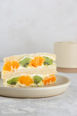 Half sandwiches bread stuffed with mascarpone cream cheese, kiwi, orange and cup of tea on a table. Homemade Korean dessert. Vertical orientation.