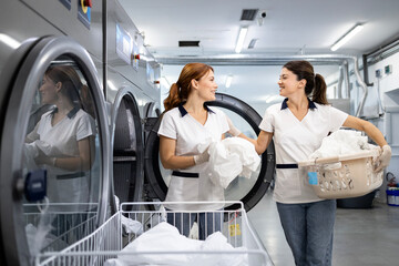 Friendly female laundry workers sorting out clothing and linen for washing in dry cleaner service. In background washing and drying machines.