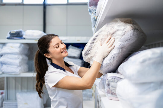 Portrait of female laundry worker standing in dry cleaning service storage room. In background, clean folded laundry ready for customer delivery.