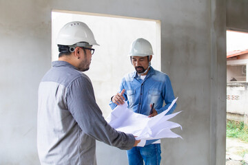 Two construction engineers in hard hats serious discussion over blueprints at construction site, Asian architect talking to foreman standing at unfinished building structure in real estate investment